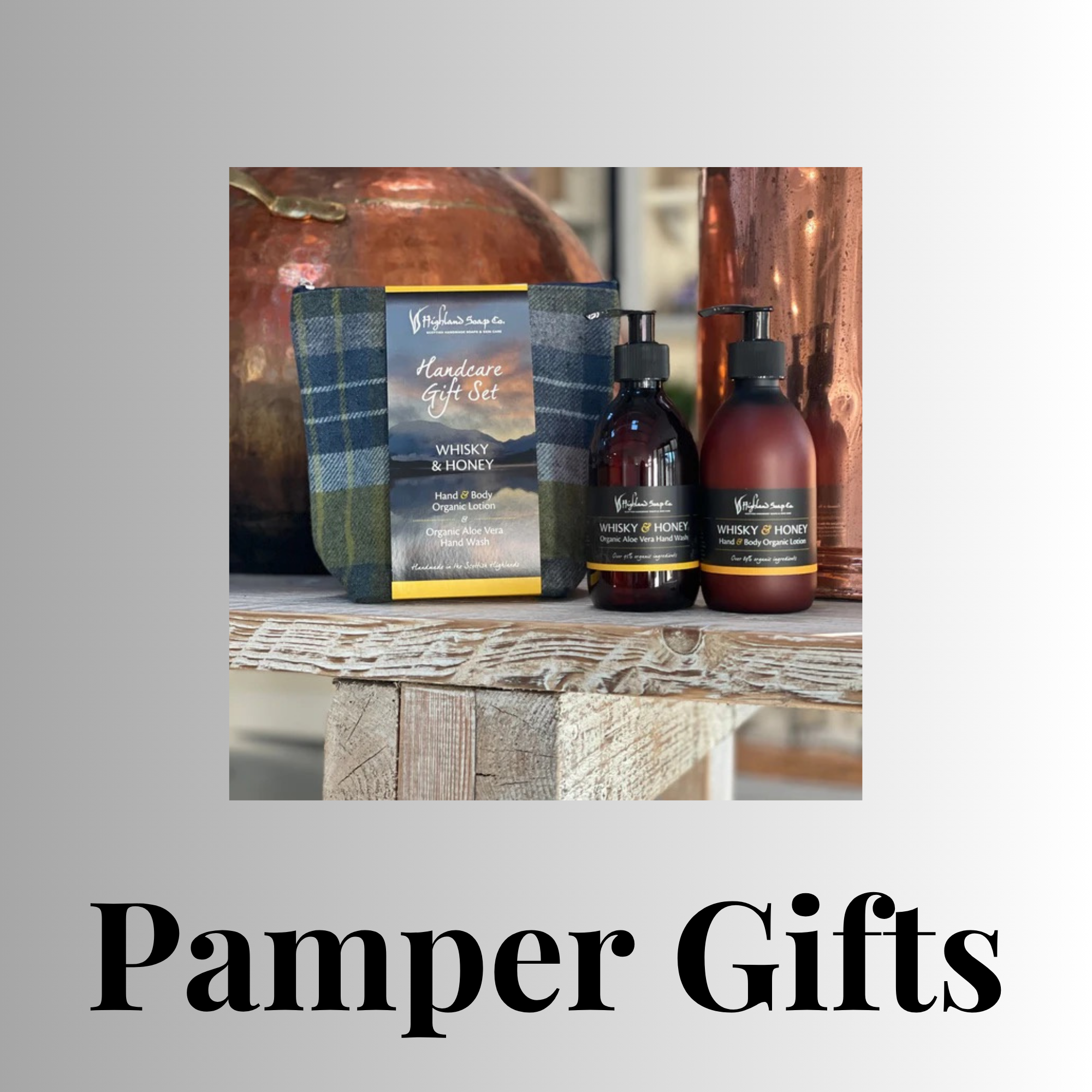 Highland Soap Co. ‘Whisky & Honey’ pamper gift set—two amber pump bottles and a blue-green tartan toiletry bag—displayed on a rustic wooden shelf, with copper stills blurred in the background. Large heading ‘Pamper Gifts’ beneath the image.