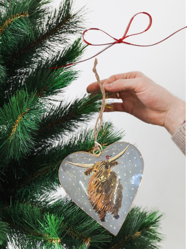 Tin heart-shaped Christmas decoration featuring a detailed illustration of a Highland cow alongside a friendly robin in a snowy scene.