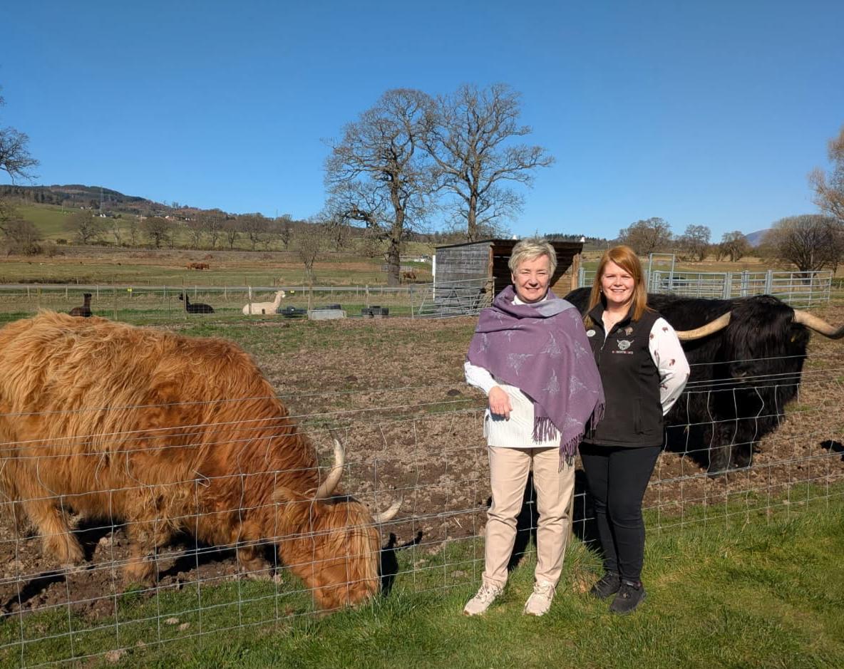 Two smiling women stand beside a fence with a grazing ginger Highland cow in front and a black Highland bull behind, sunny Beauly fields in background.
