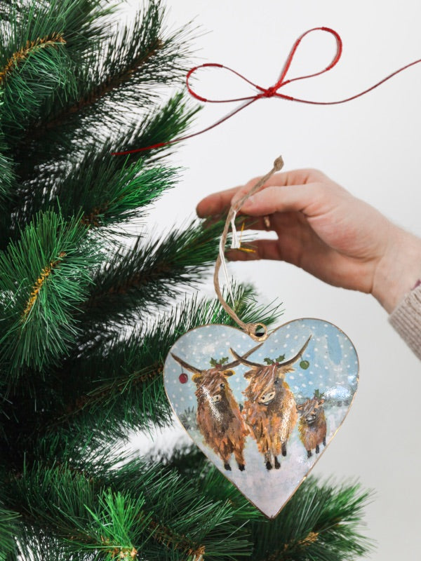 Tin heart-shaped hanging Christmas decoration depicting three adorable Highland cows adorned with Christmas baubles, holly, and festive details.