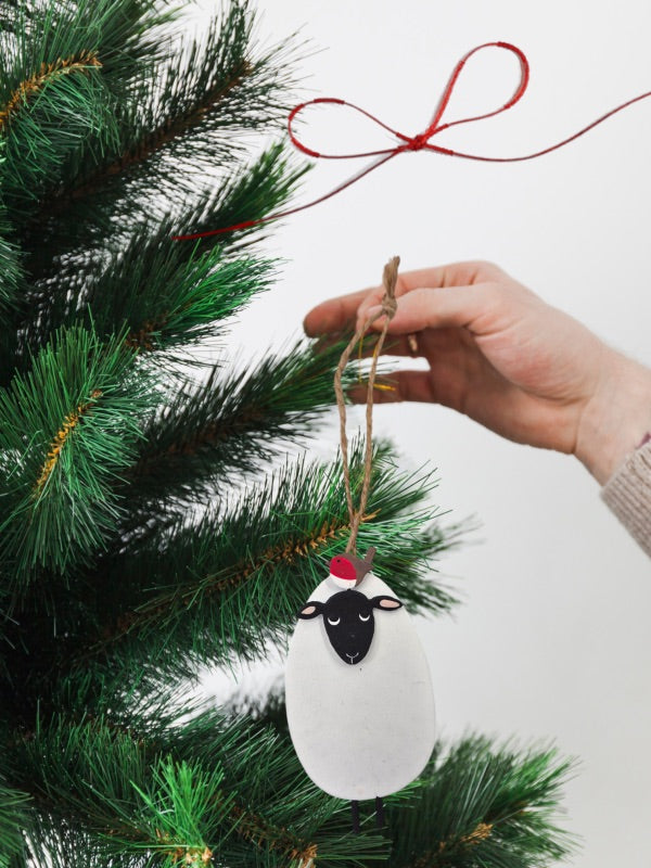 Hanging Christmas tree ornament depicting a black-faced sheep with a small robin on its head, held near a green pine branch.