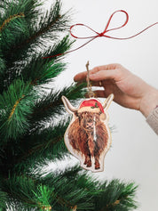 Tin heart Christmas decoration featuring a charming Highland cow wearing a Santa hat and surrounded by festive holly sprigs.
