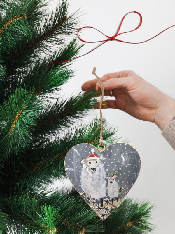 Tin heart Christmas decoration illustrating a cozy scene with a sheep wearing a Santa hat alongside a lamb accented with a sprig of holly, perfect for festive décor.